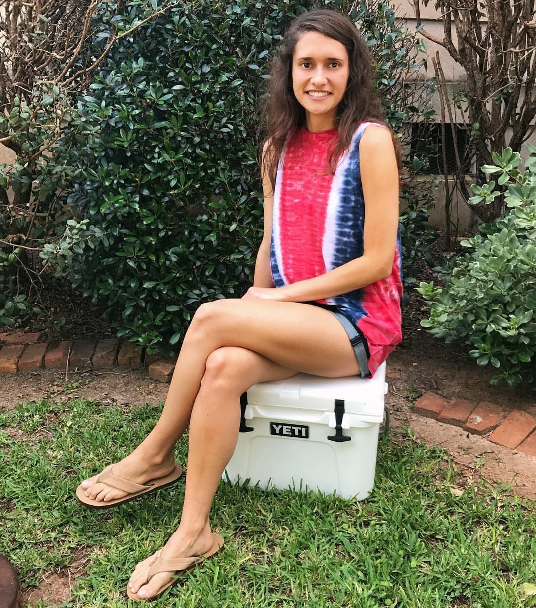 Smiling woman in a red and blue tie-dye top sitting on a white cooler outdoors.
