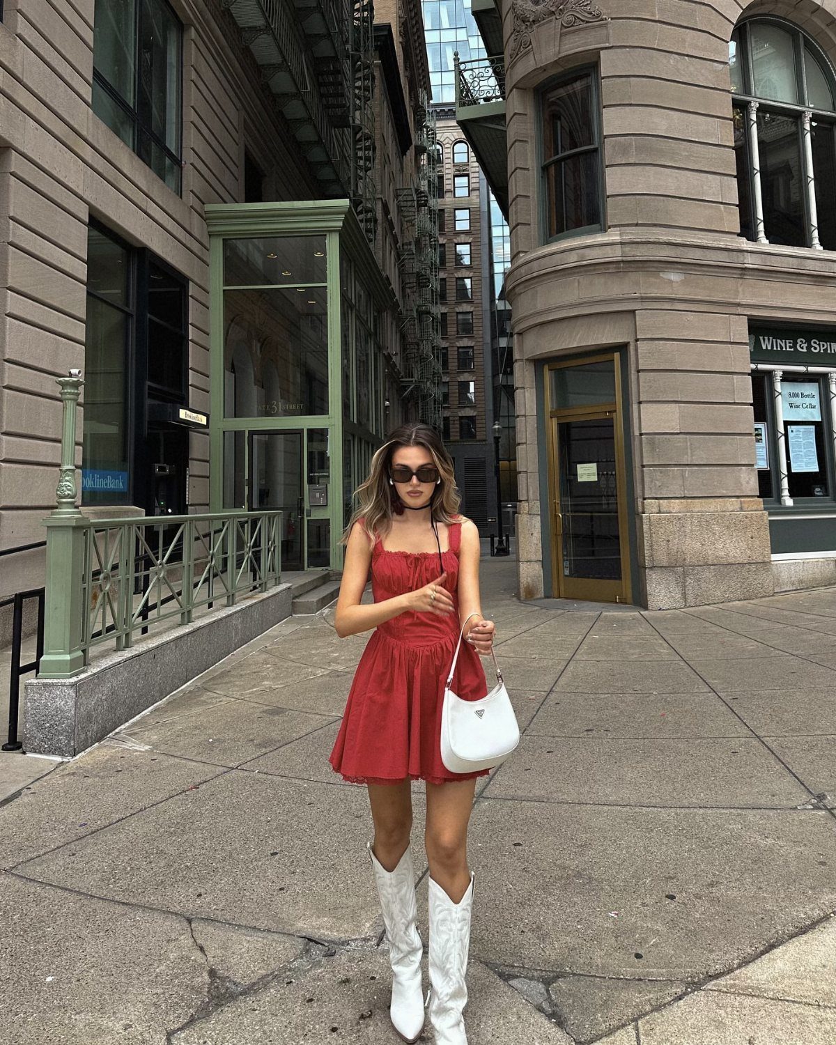 Woman in a red mini dress and white cowboy boots standing on a city street.