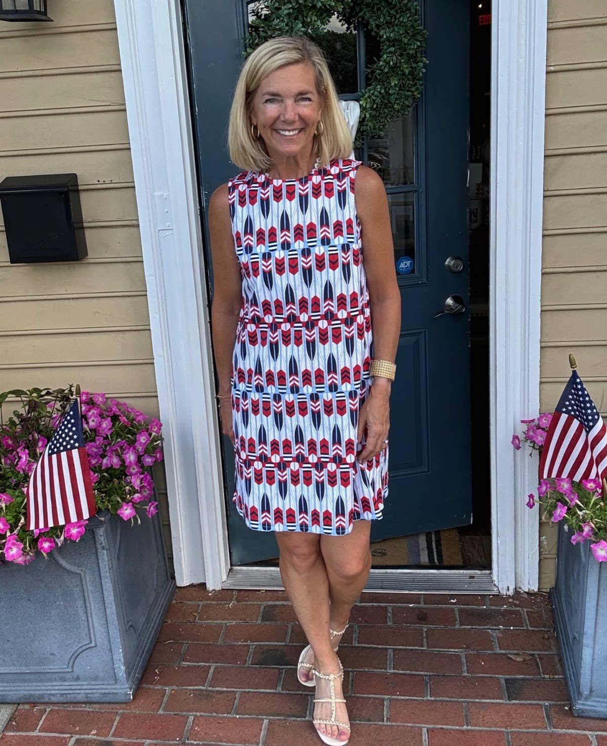 A blonde woman smiling in a patterned sleeveless dress and beige sandals standing by a doorway.