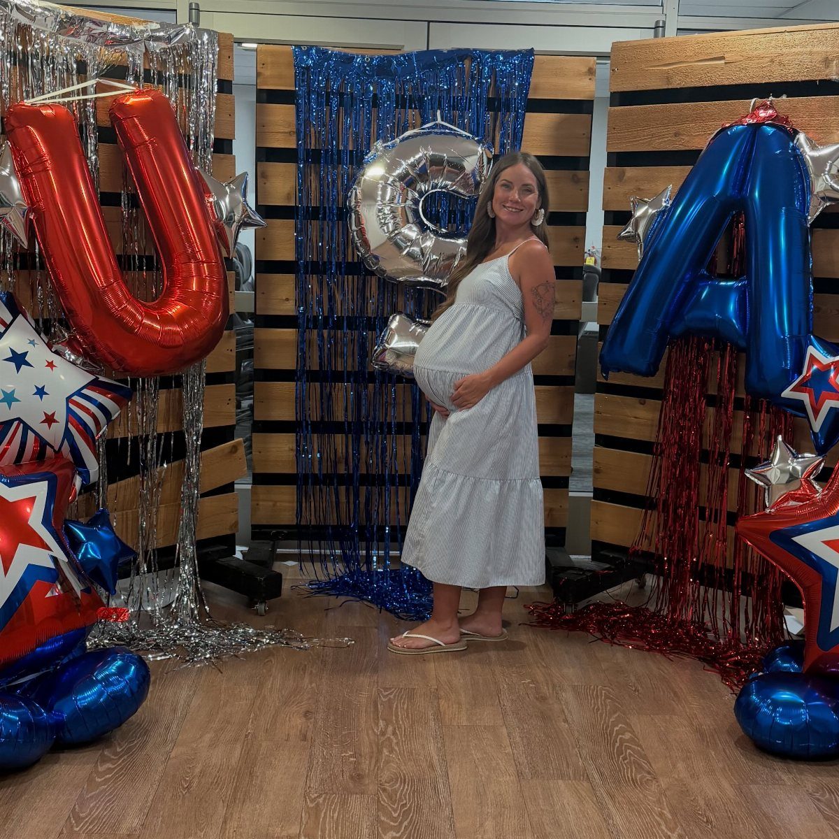 A pregnant woman wearing a white and navy striped dress standing among patriotic balloons.