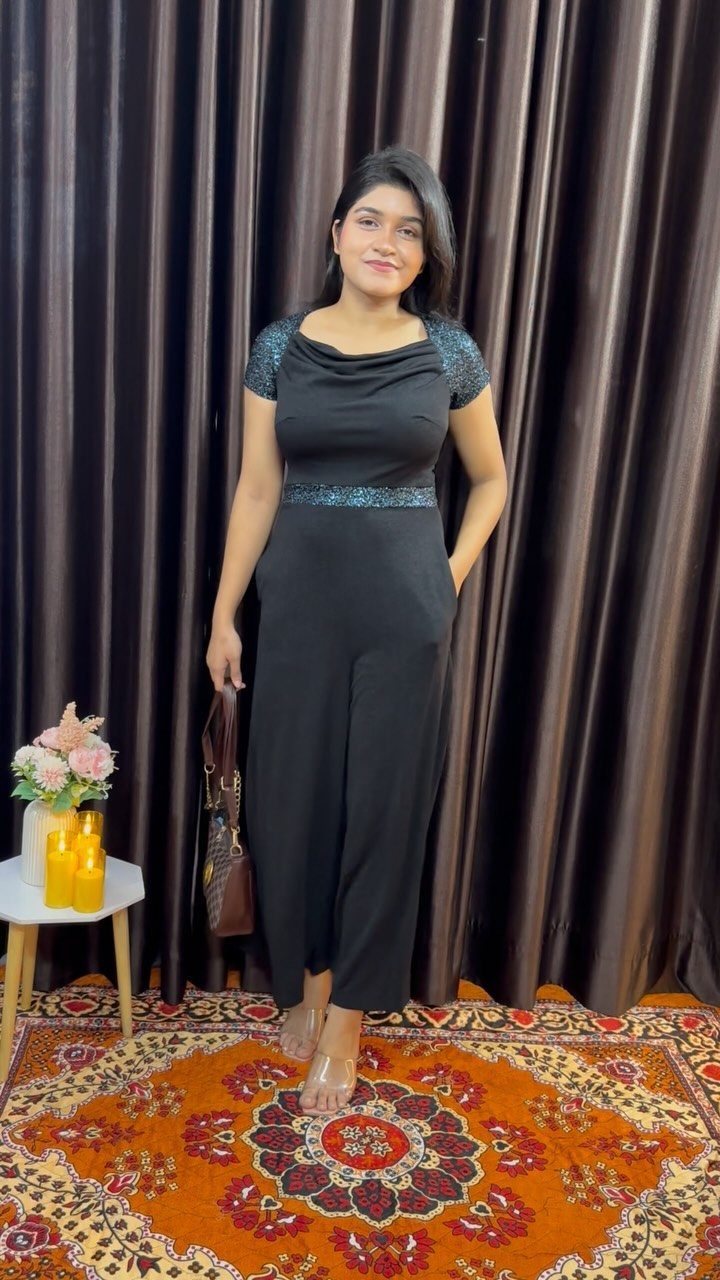 Woman wearing a black sequined jumpsuit standing in front of dark brown curtains.