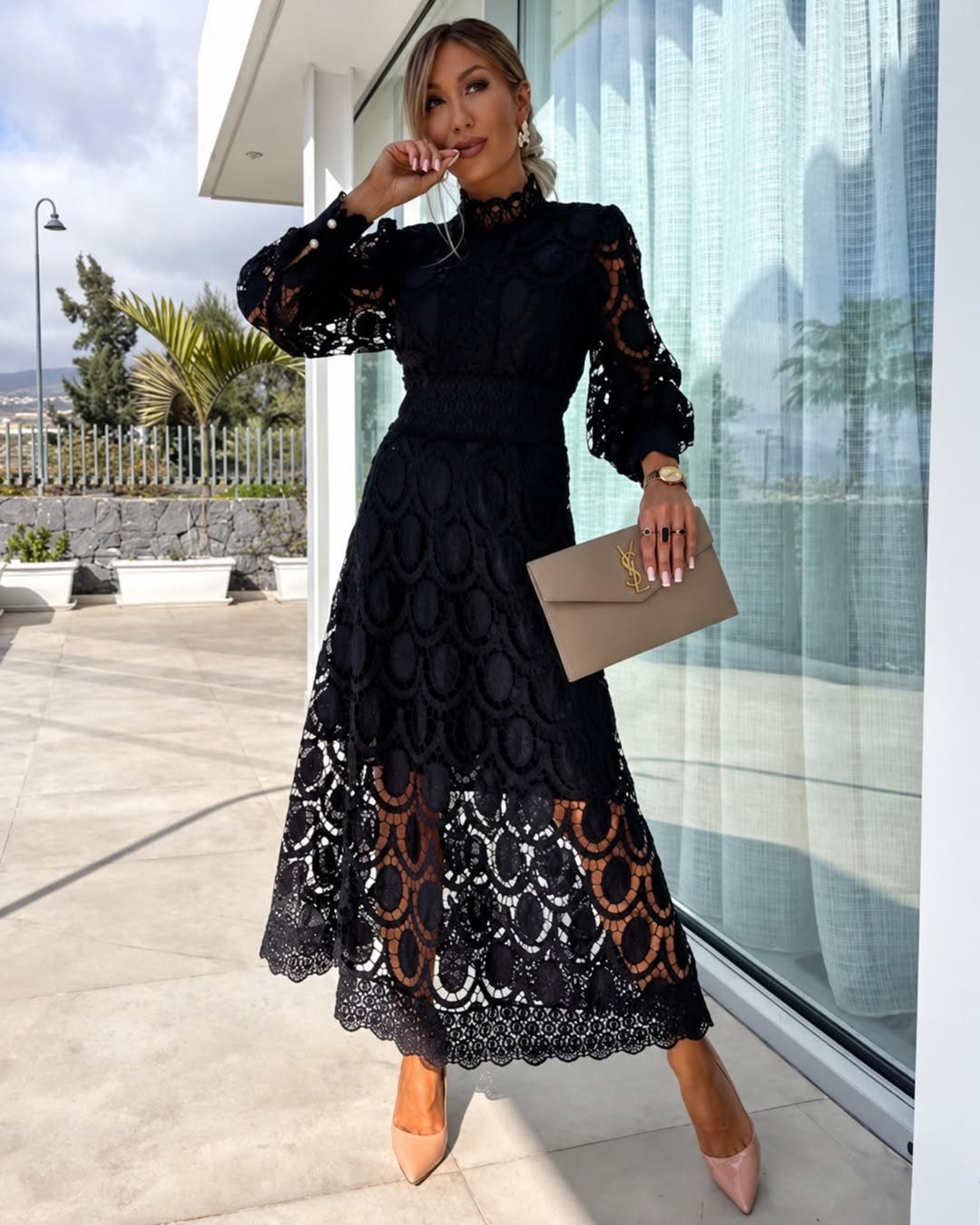 A woman standing on a terrace wearing an elegant long-sleeved black lace midi dress and nude heels.