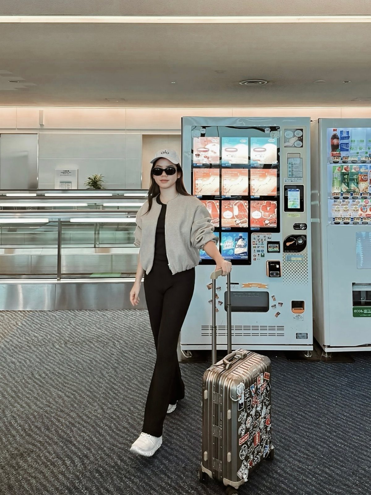 A woman in a grey bomber jacket and black jumpsuit walks with a suitcase through an airport.