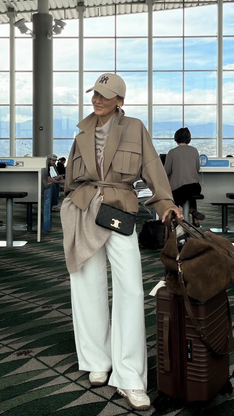 A woman in a beige jacket and white wide-leg trousers standing in an airport terminal.