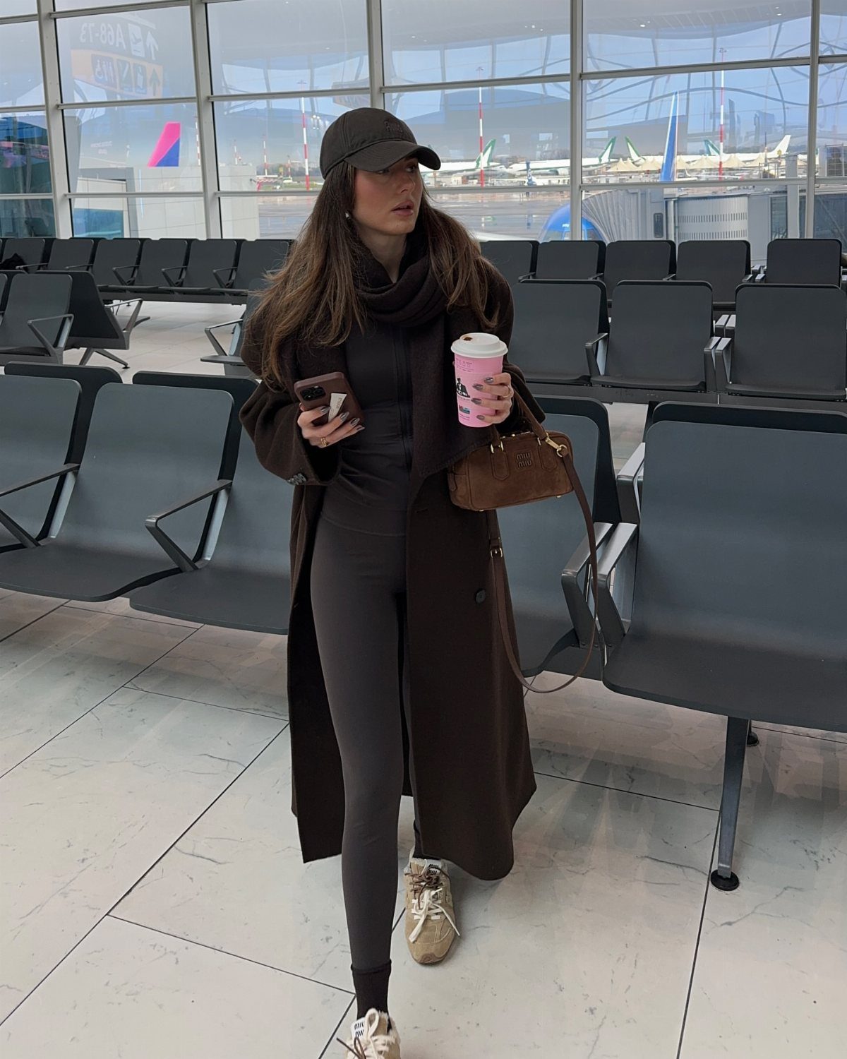A woman in a long brown coat and leggings walks through an airport terminal holding coffee.
