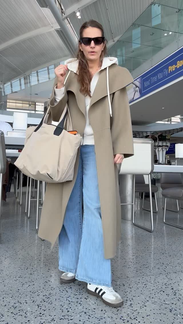 Woman in a long beige coat and wide-leg blue jeans standing in an airport terminal.