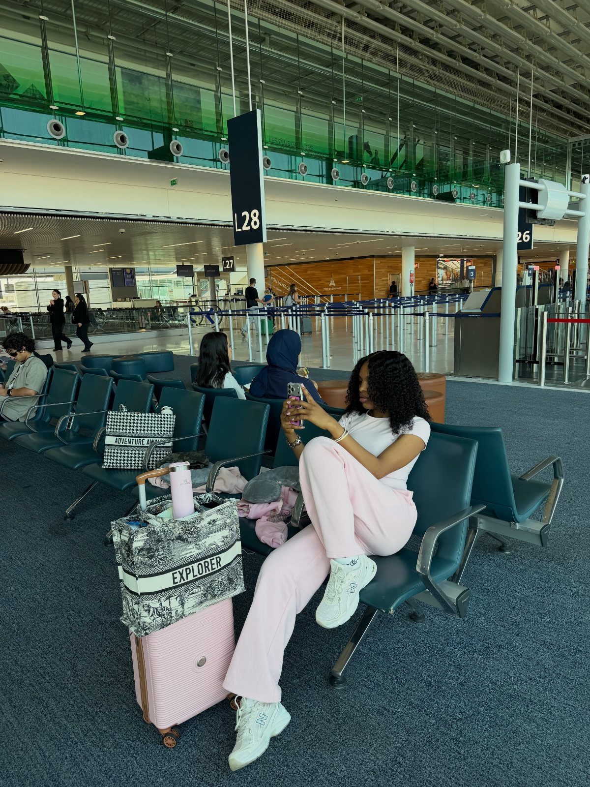 A woman in pink clothing sits in an airport terminal next to a large patterned tote bag.