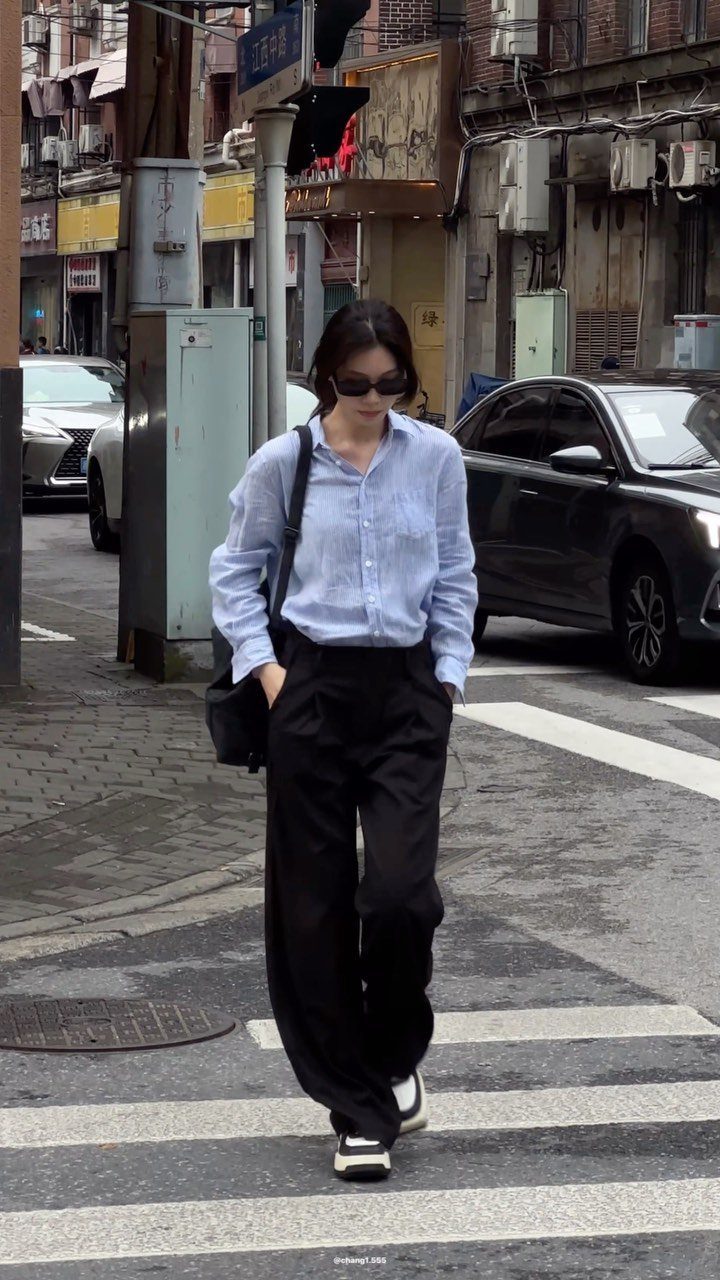 A woman walking down a city street wearing black wide-leg trousers and a blue shirt.