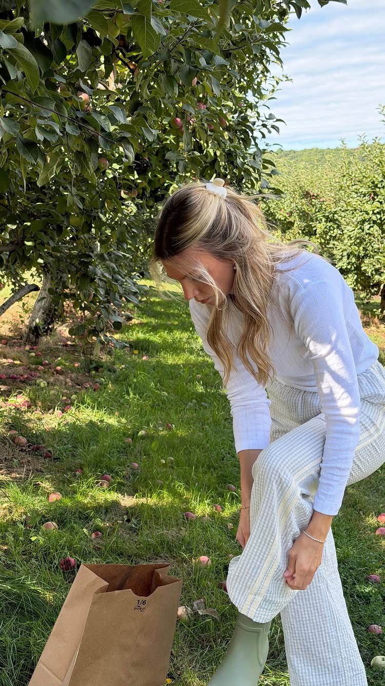A woman wearing a white ribbed top and striped pants bends down in an apple orchard.