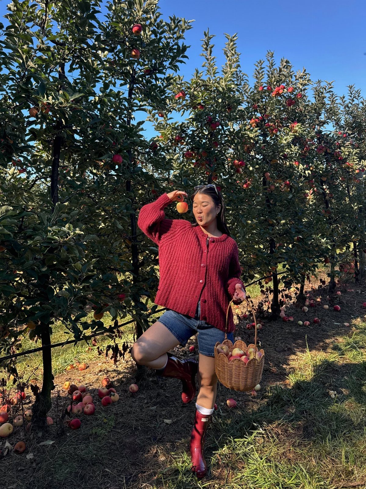 Woman in a red cardigan and denim shorts wearing tall red boots in an apple orchard.
