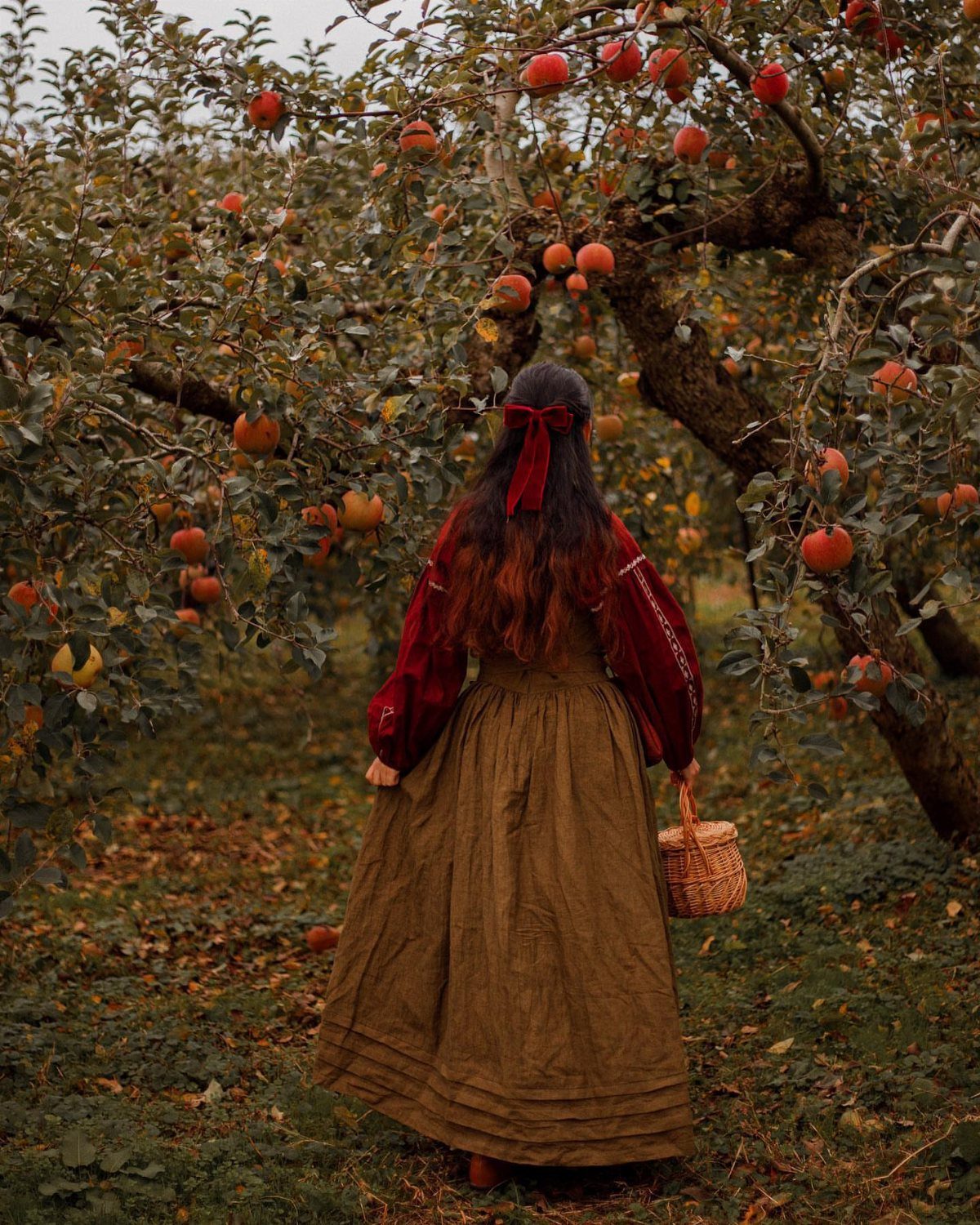 Woman in a long olive skirt and red blouse walking through an apple orchard.