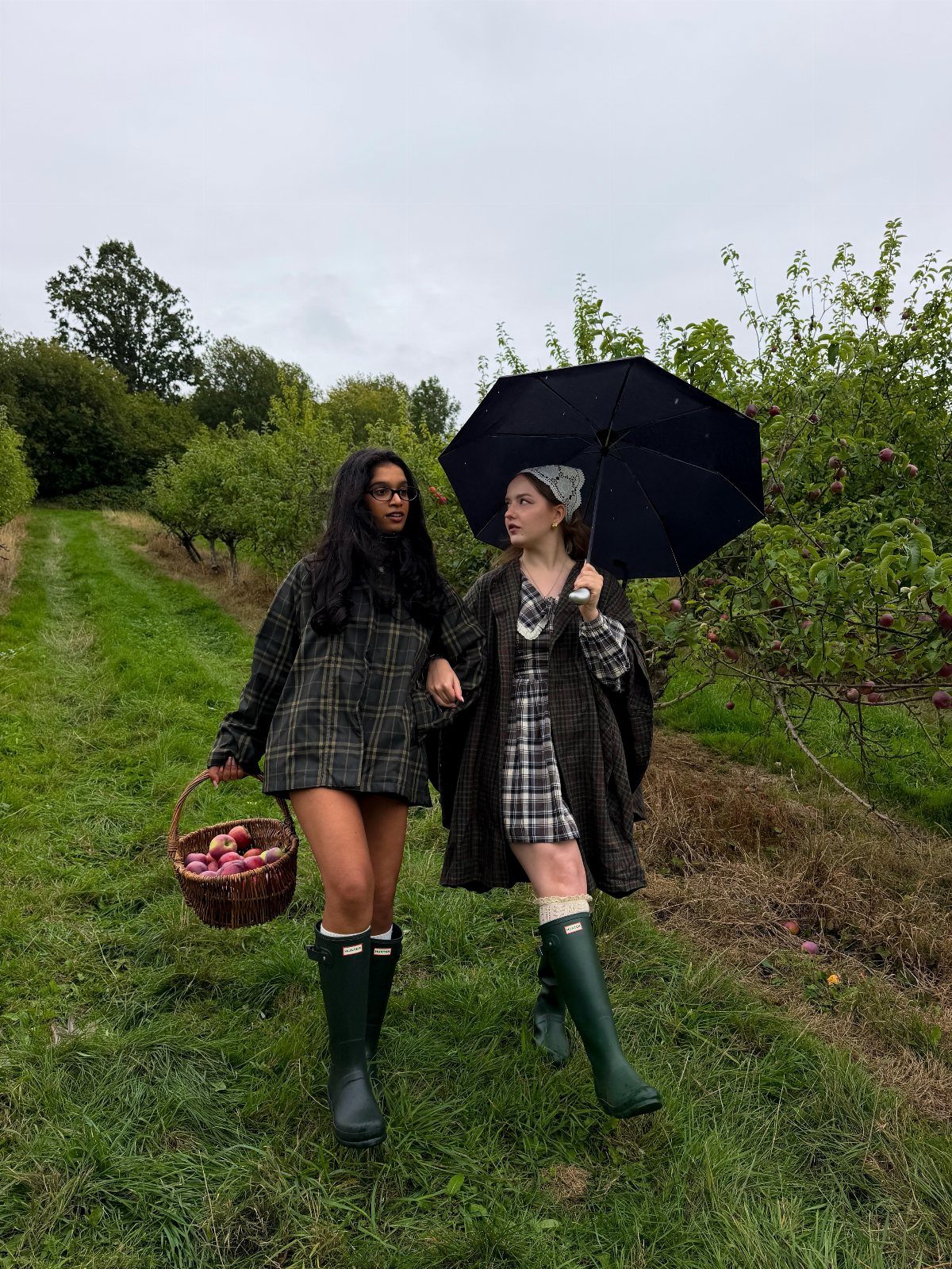 Two women walking through an apple orchard wearing plaid clothing and green rain boots.
