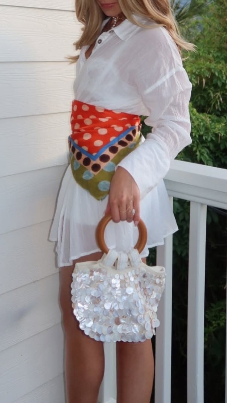 Woman in a white shirt and skirt wearing a colorful waist scarf and holding a sequined bag.