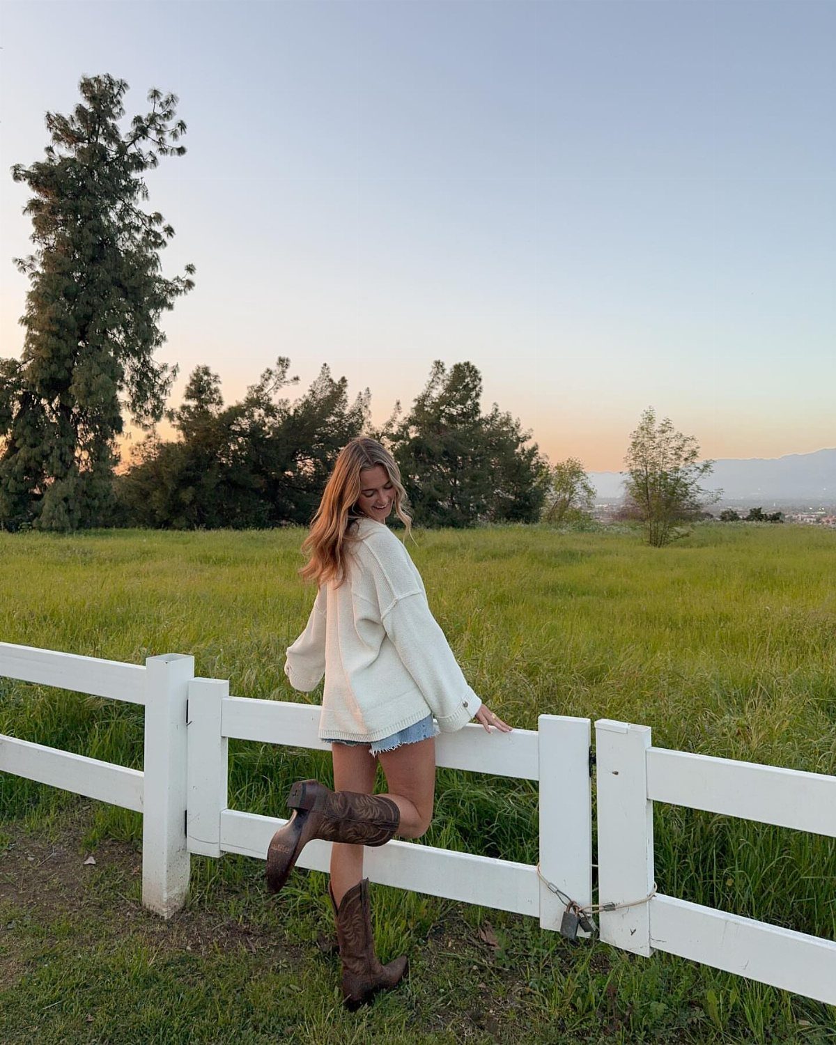 Woman in white sweater and denim shorts wearing brown cowgirl boots by a white fence.