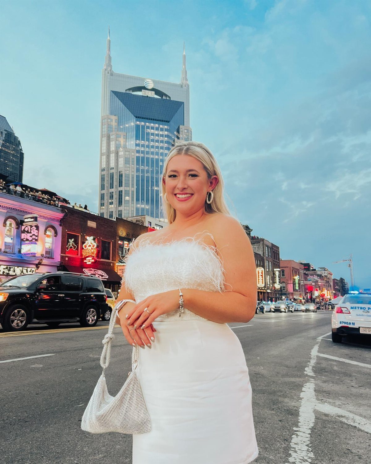 Smiling blonde woman wearing a white feathered dress and holding a silver handbag on a city street.