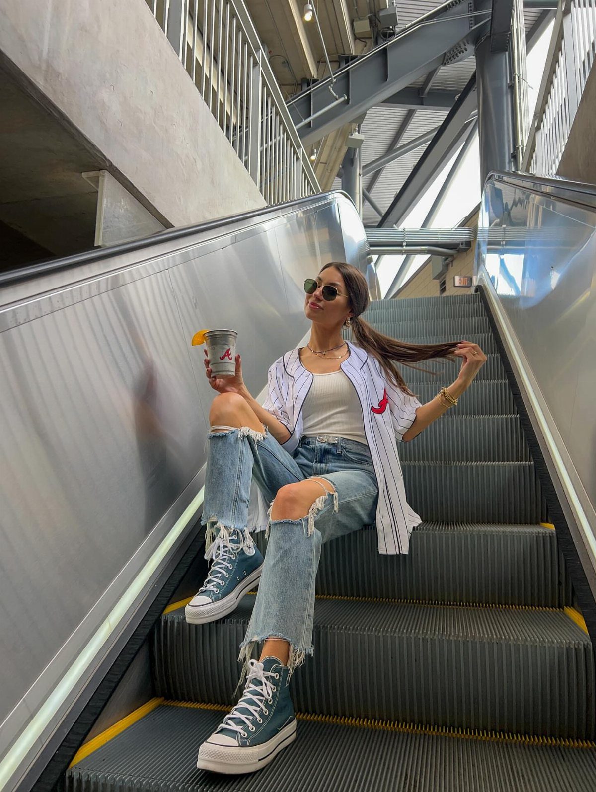 Woman in ripped jeans and a baseball jersey sitting on an escalator wearing blue high-top sneakers.