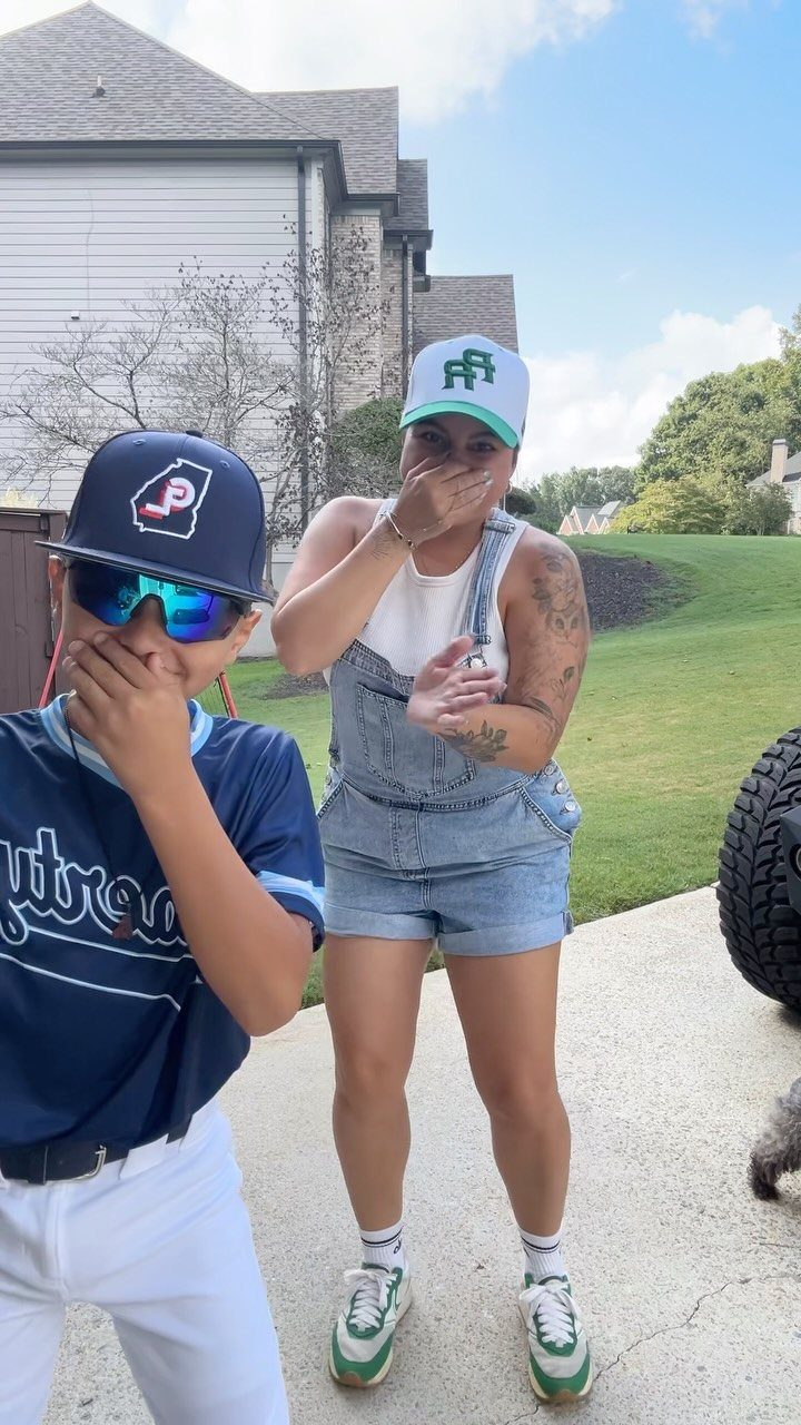 A child in a baseball jersey and a woman in denim overalls posing outdoors.