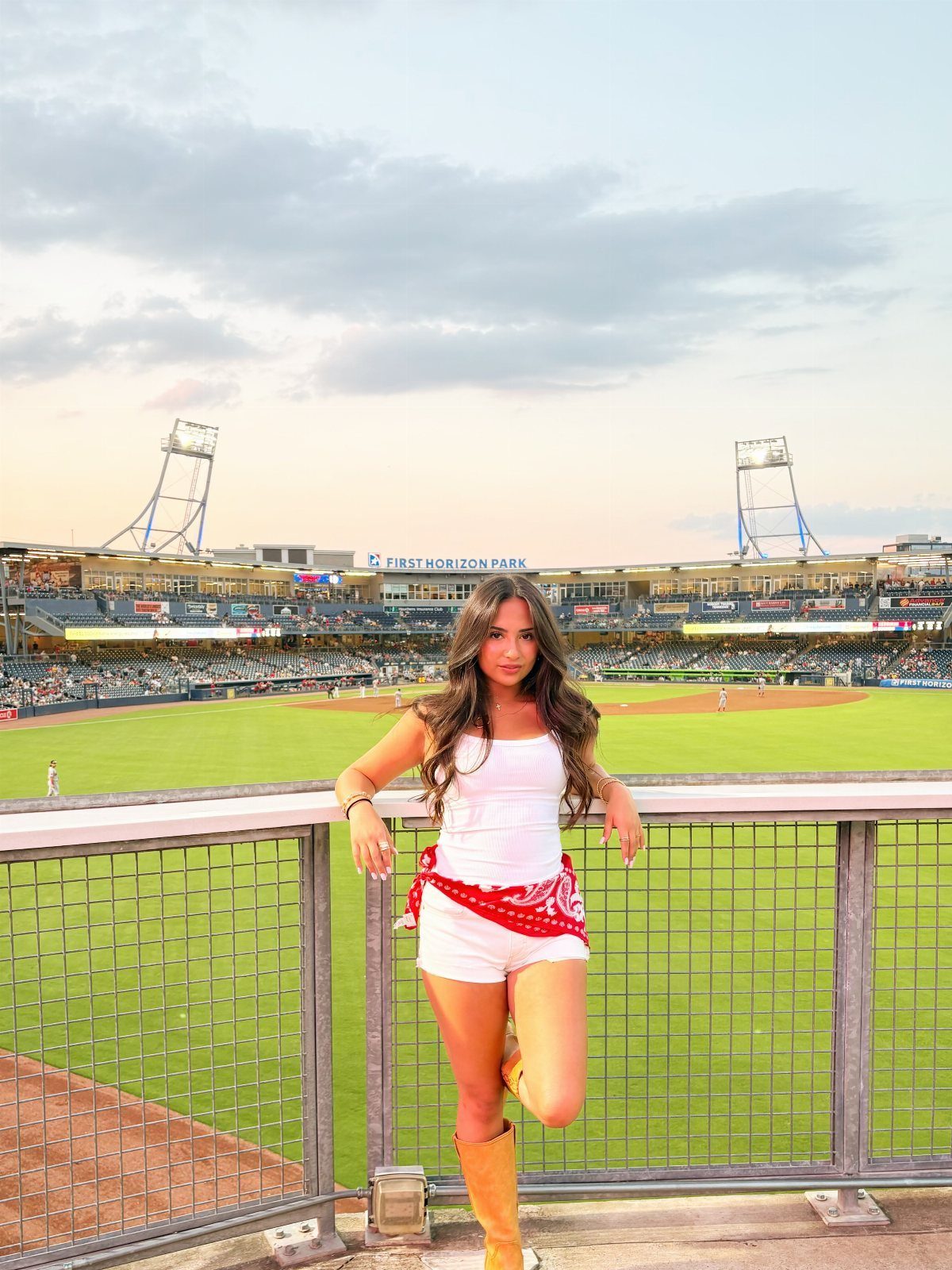 Woman in a white tank top and shorts posing at a baseball stadium during sunset.