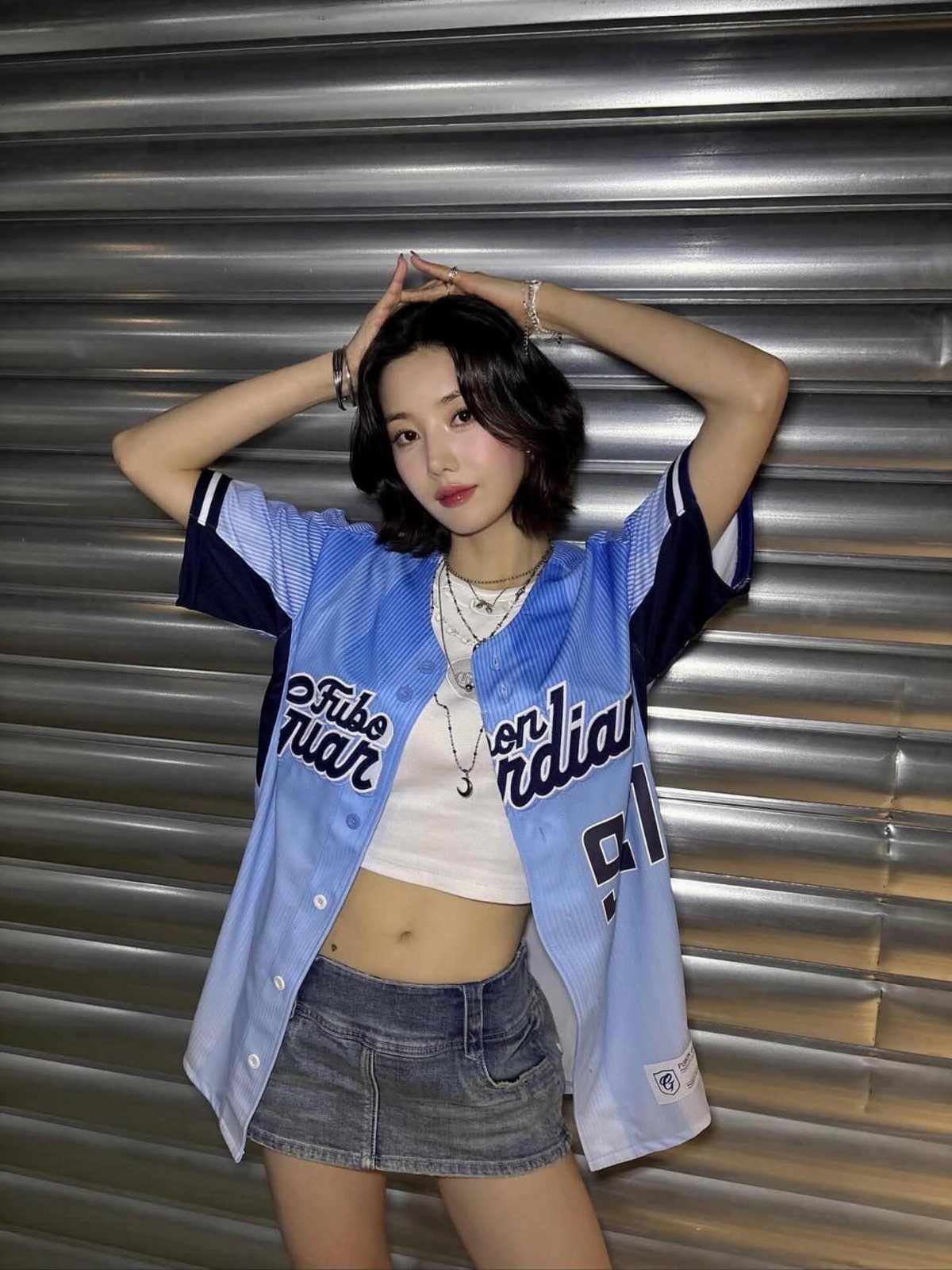 Young woman wearing an open light blue baseball jersey over a white crop top and denim skirt.