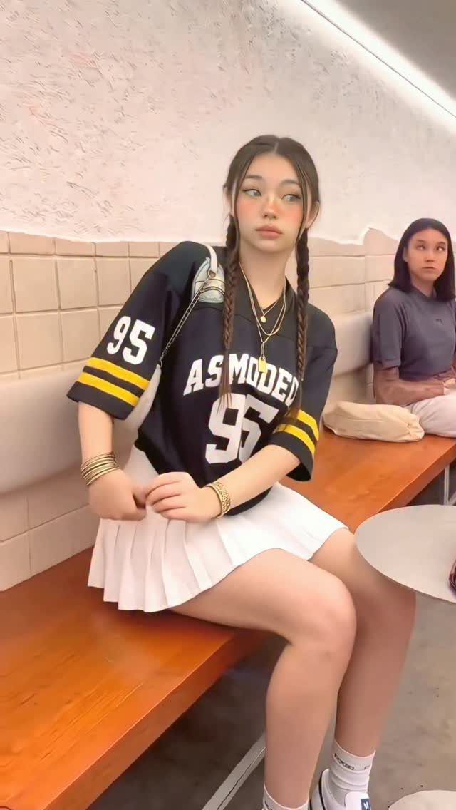 Young woman sitting on a bench wearing a black baseball jersey and white pleated skirt.
