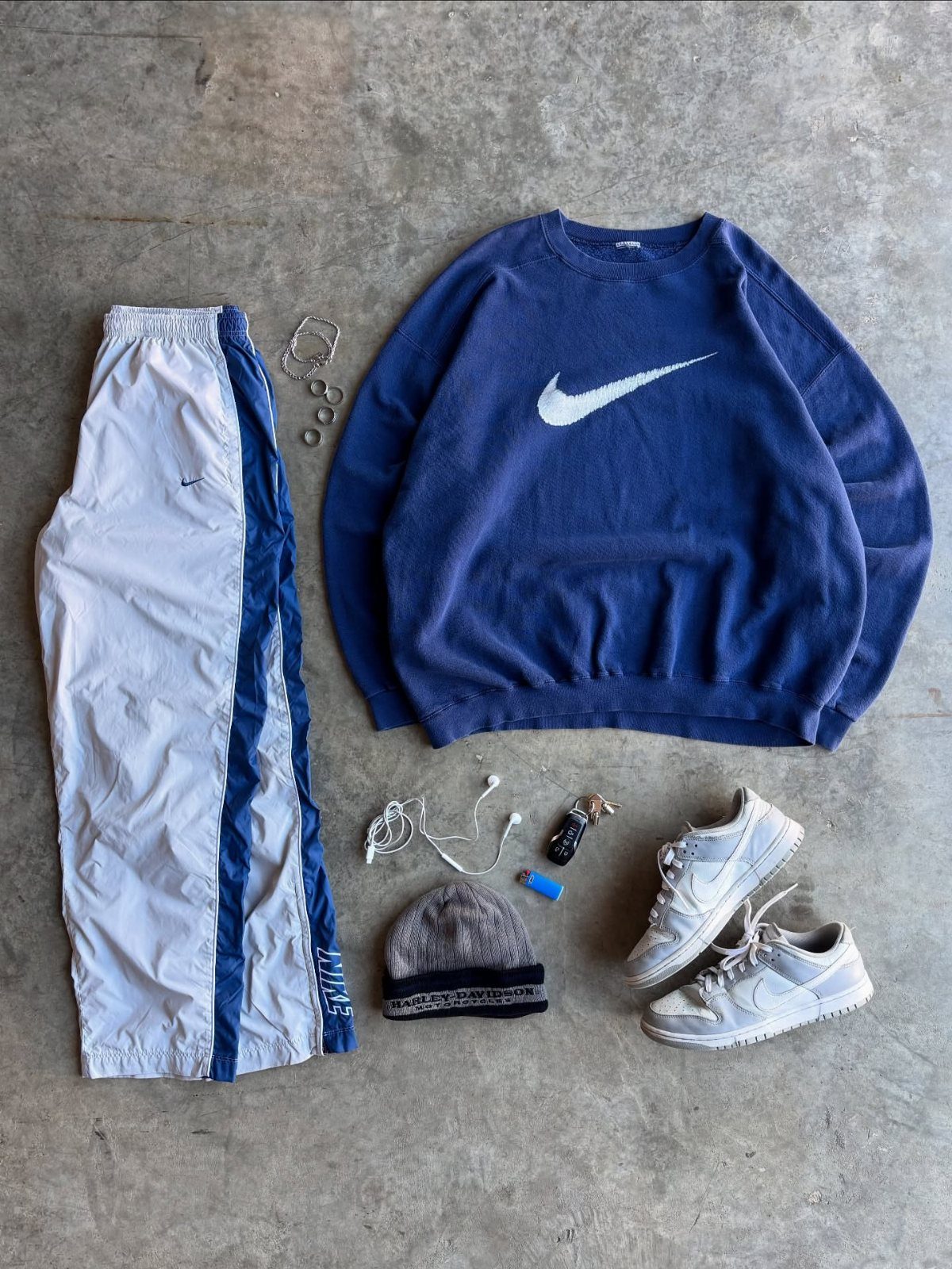 White and blue track pants, a navy Nike sweatshirt, and white sneakers laid on concrete.