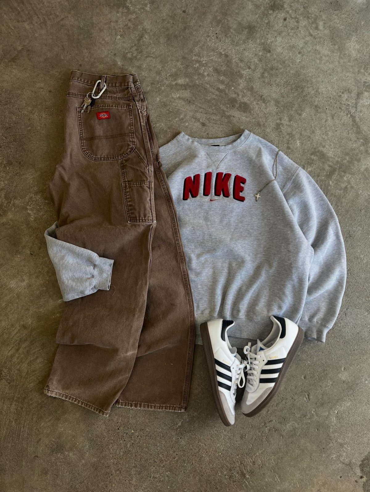 Grey Nike sweatshirt, brown carpenter pants, and white Adidas sneakers laid out on concrete.