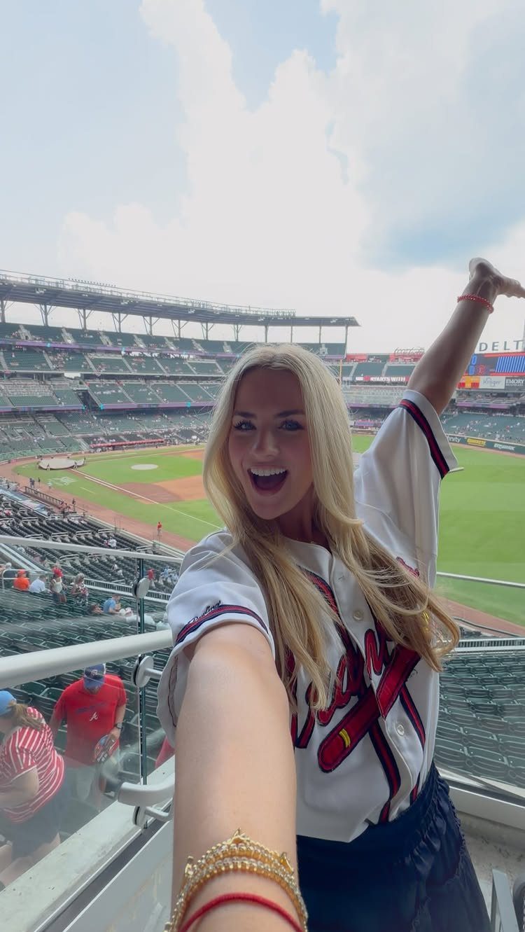 Blonde woman smiling in a white baseball jersey at a crowded stadium.