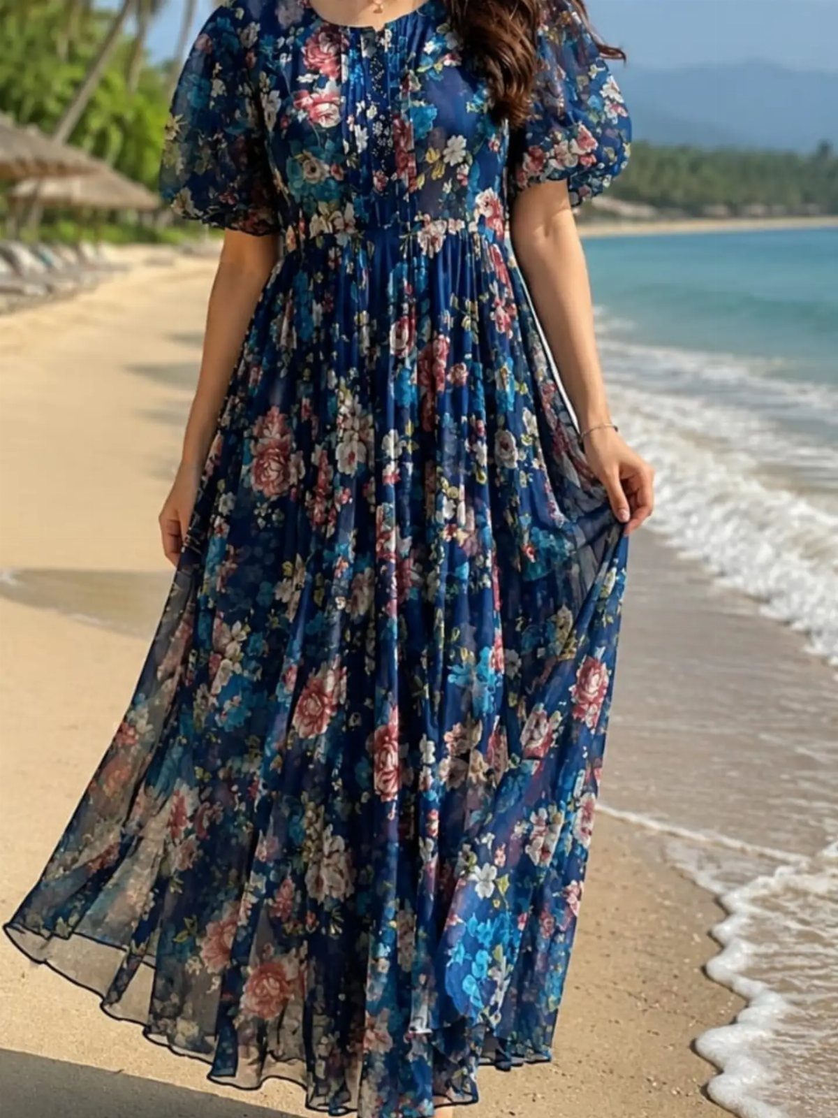 Woman wearing a blue floral maxi dress standing on a sandy beach by the ocean.