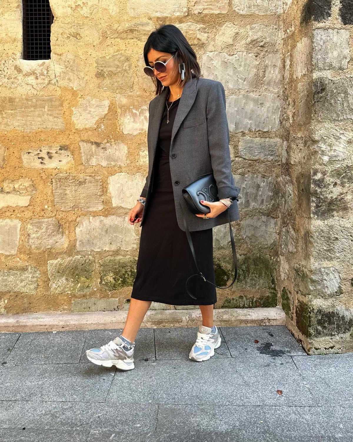 Woman in a long grey blazer and black dress walking past a stone wall.