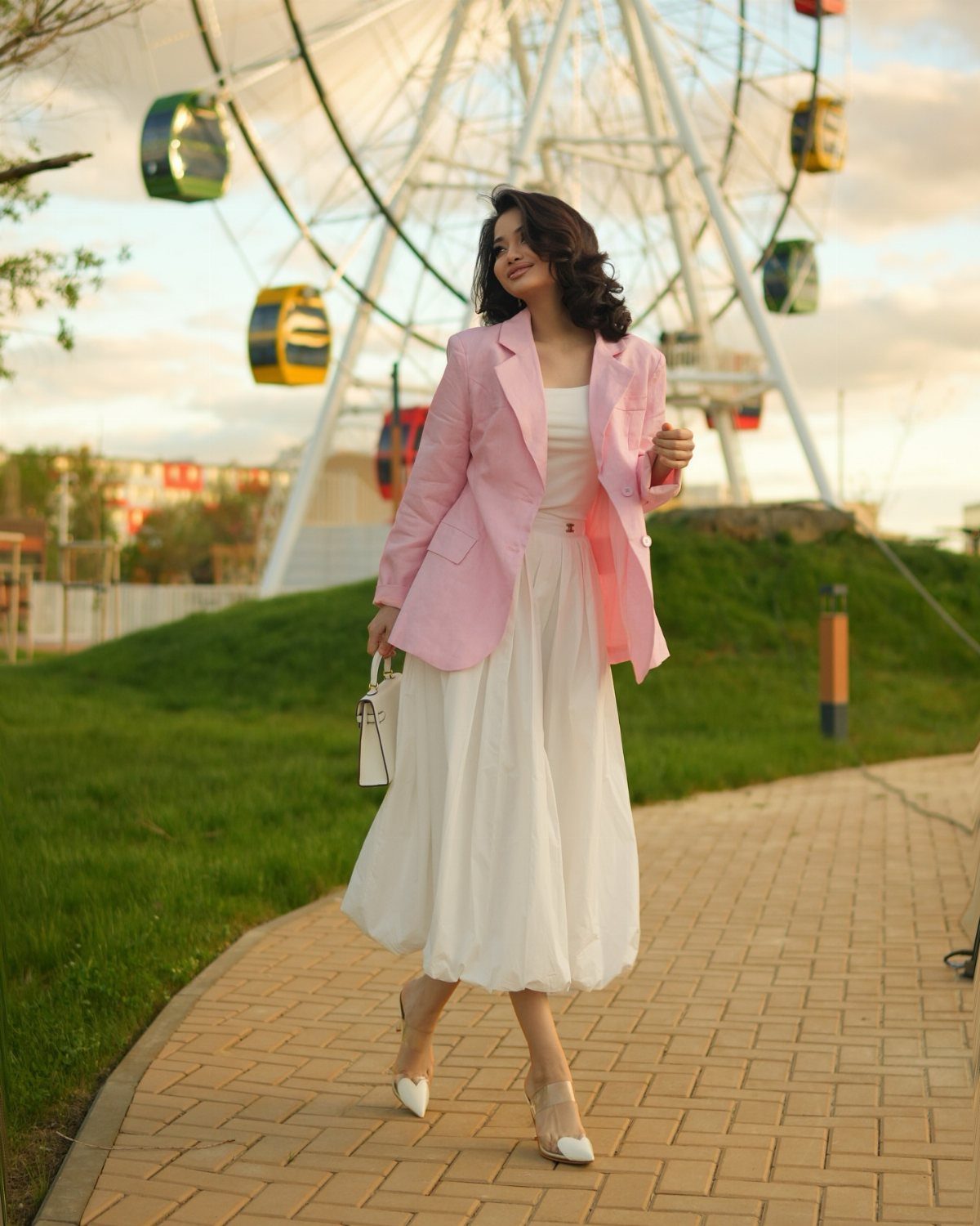 Woman in a pink blazer and white maxi dress walking near a Ferris wheel.
