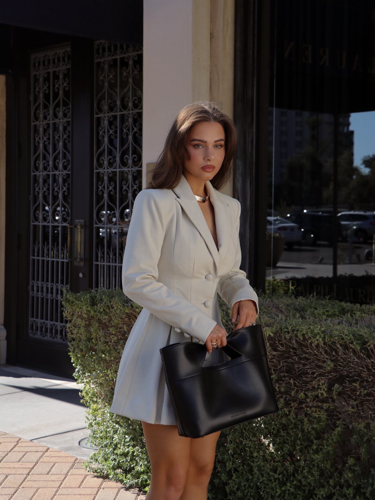 Woman in a white blazer and matching flared dress holding a black handbag.