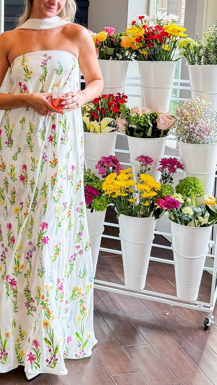 Woman in a white strapless maxi dress with a colorful floral pattern standing by flowers.