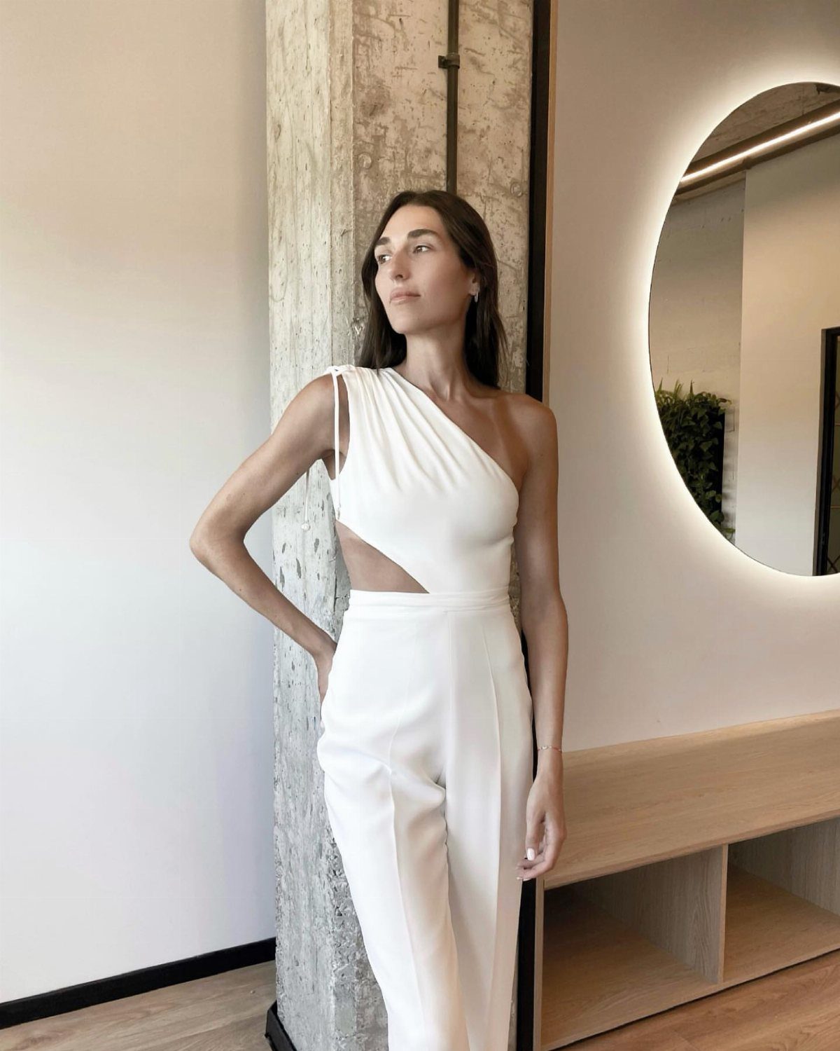 Woman in a white one-shoulder jumpsuit with a side cutout standing by a pillar.