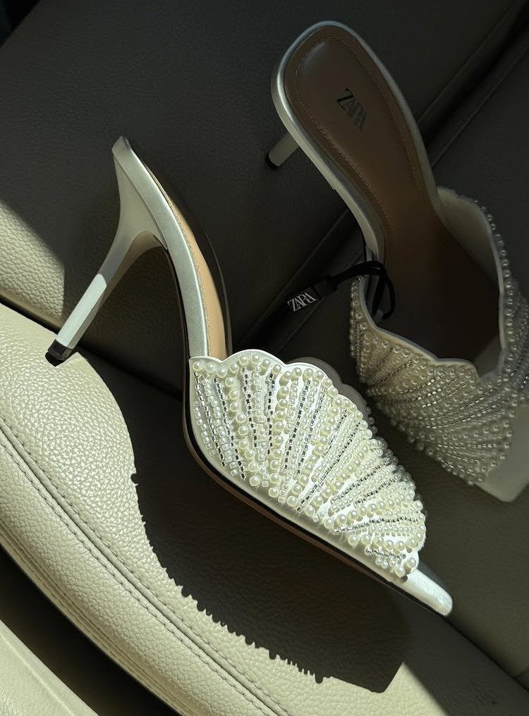 White pearl-embellished high-heeled mule sandals resting on a cream leather car seat.