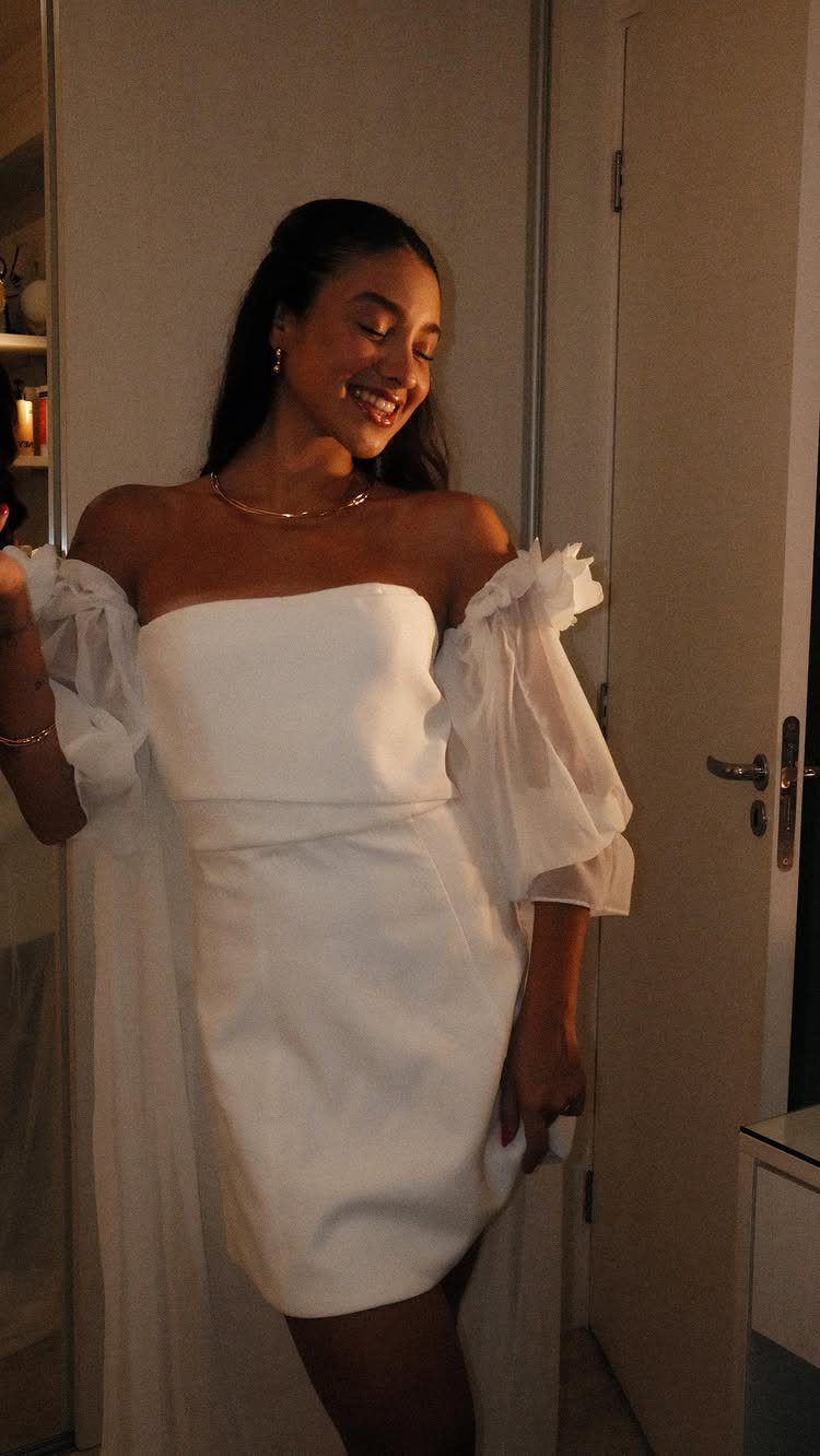 A smiling woman wearing a white strapless mini dress with large sheer puff sleeves.