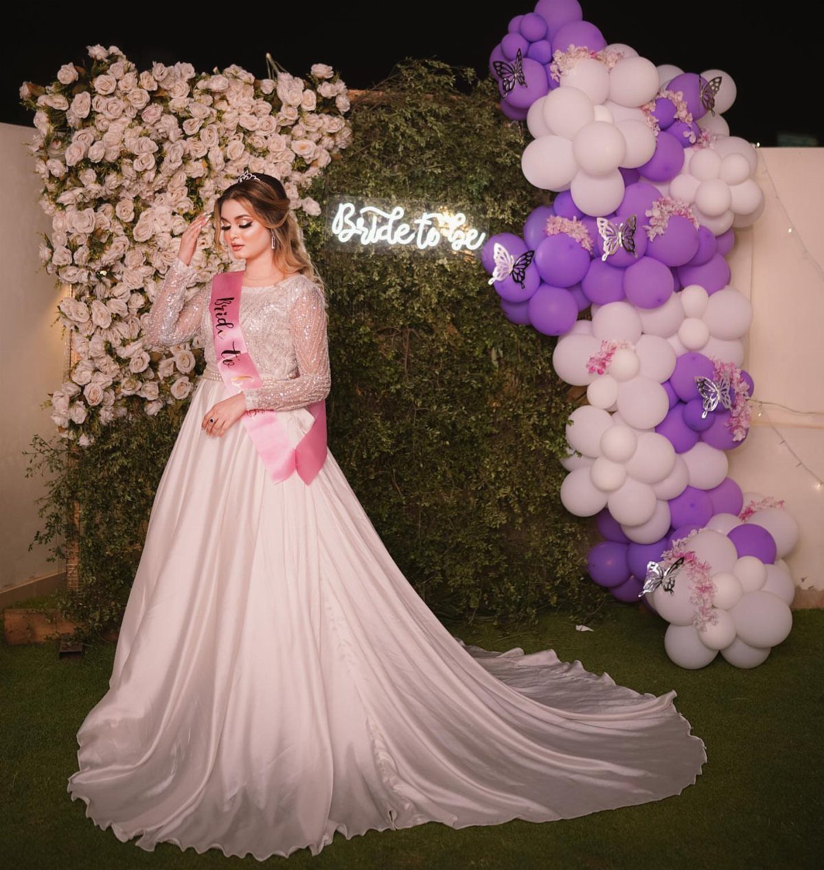 Woman in a white sequined gown and pink sash posing against a floral and balloon backdrop.