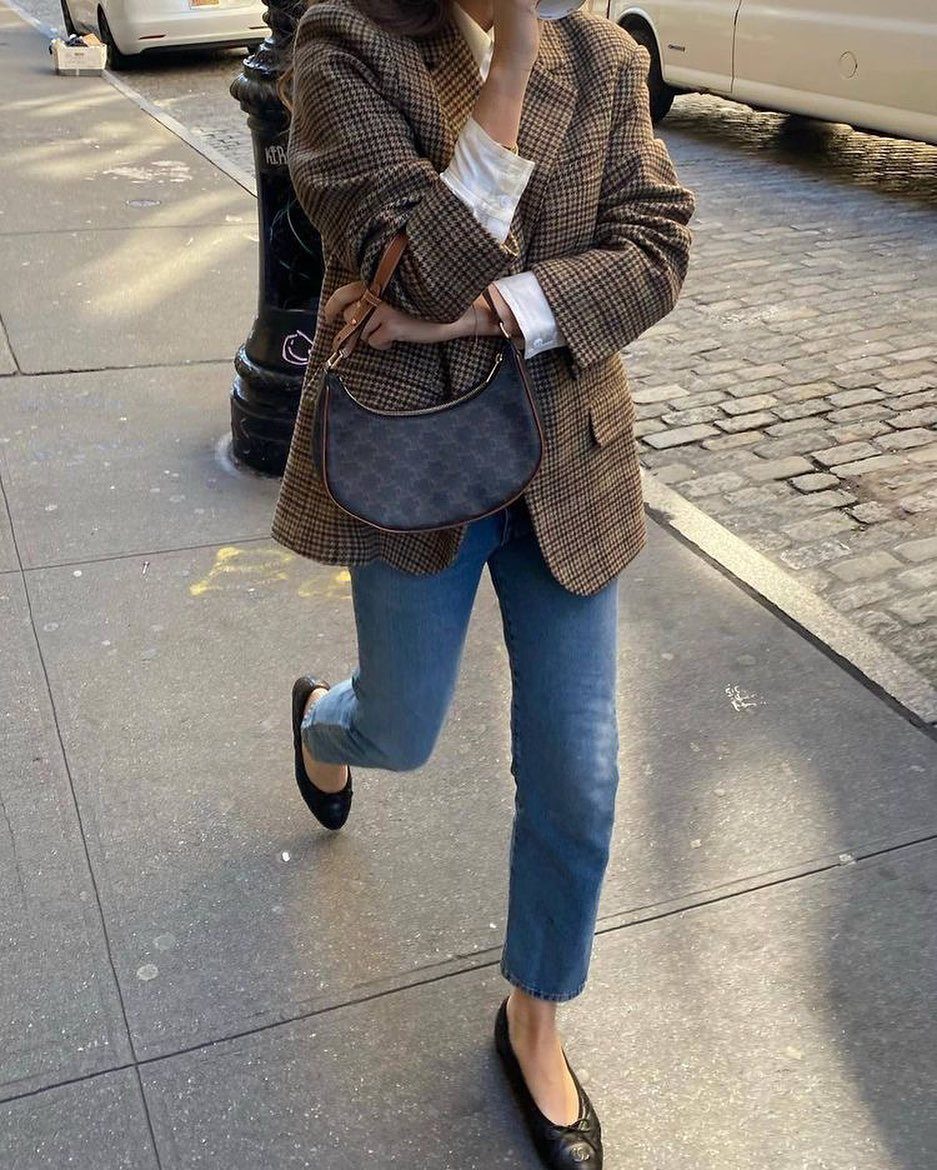 Person wearing a plaid blazer, white shirt, blue jeans, and black flats walking on a sidewalk.