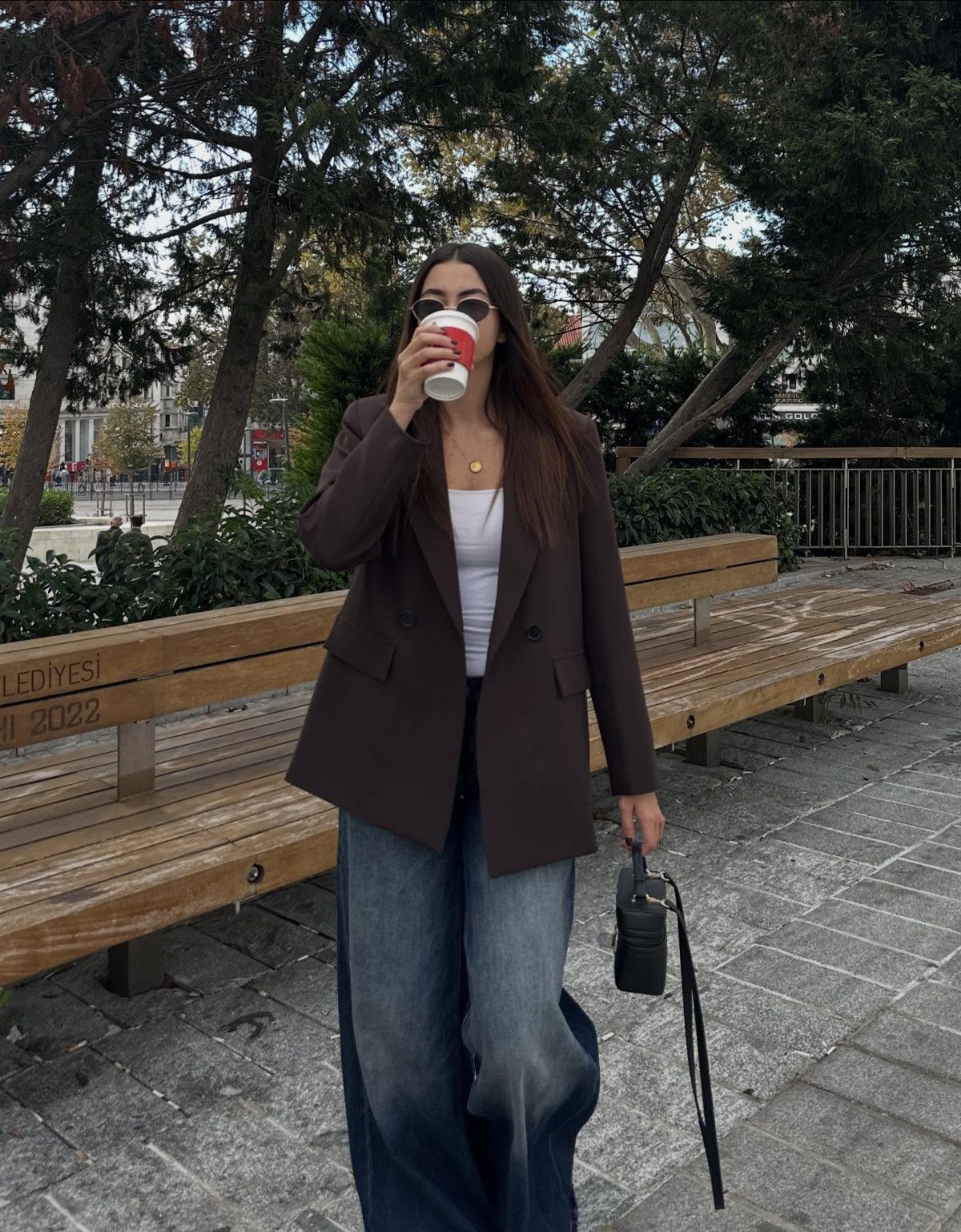 Woman wearing a dark brown oversized blazer, white top, and wide-leg denim trousers while drinking coffee.
