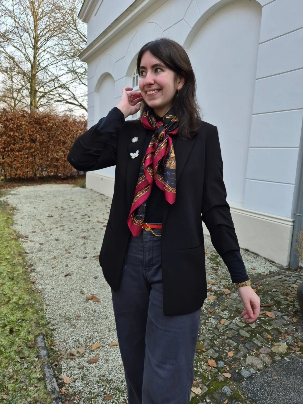 A smiling woman wearing a black blazer, patterned neck scarf, and dark grey trousers outdoors.
