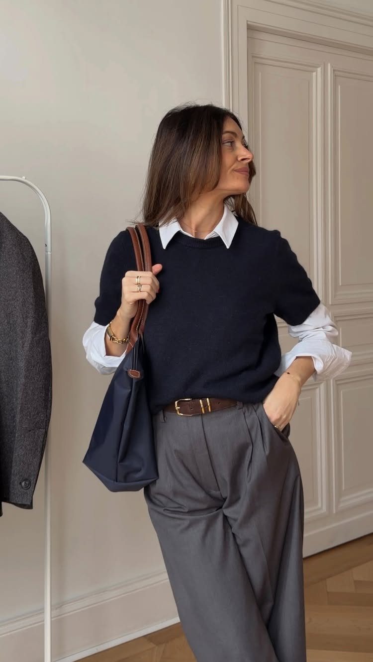Woman wearing a navy sweater, white collared shirt, and grey trousers standing indoors.