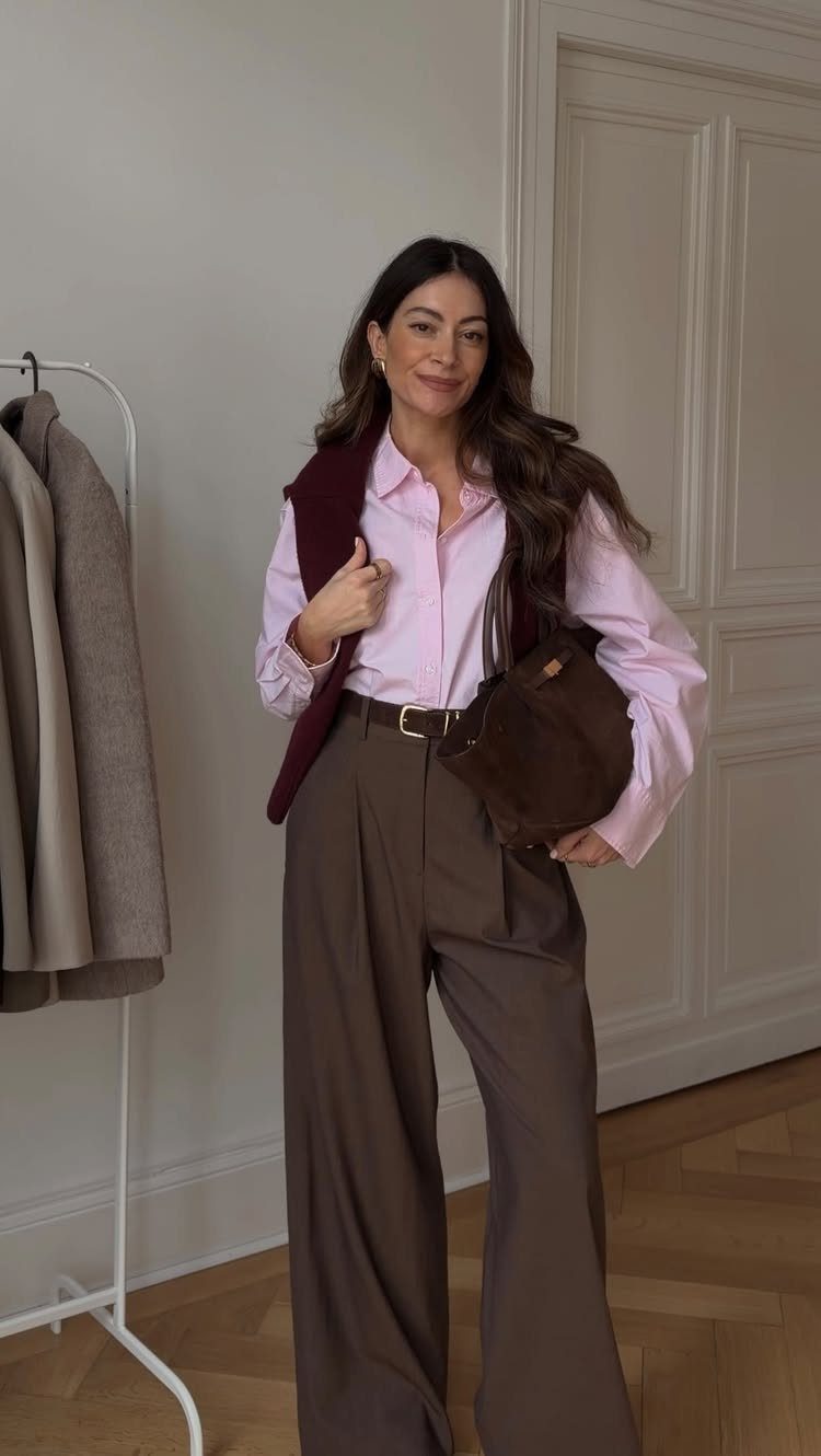 A woman smiling while wearing a pink shirt, brown wide-leg trousers, and a maroon sweater draped over her shoulders.