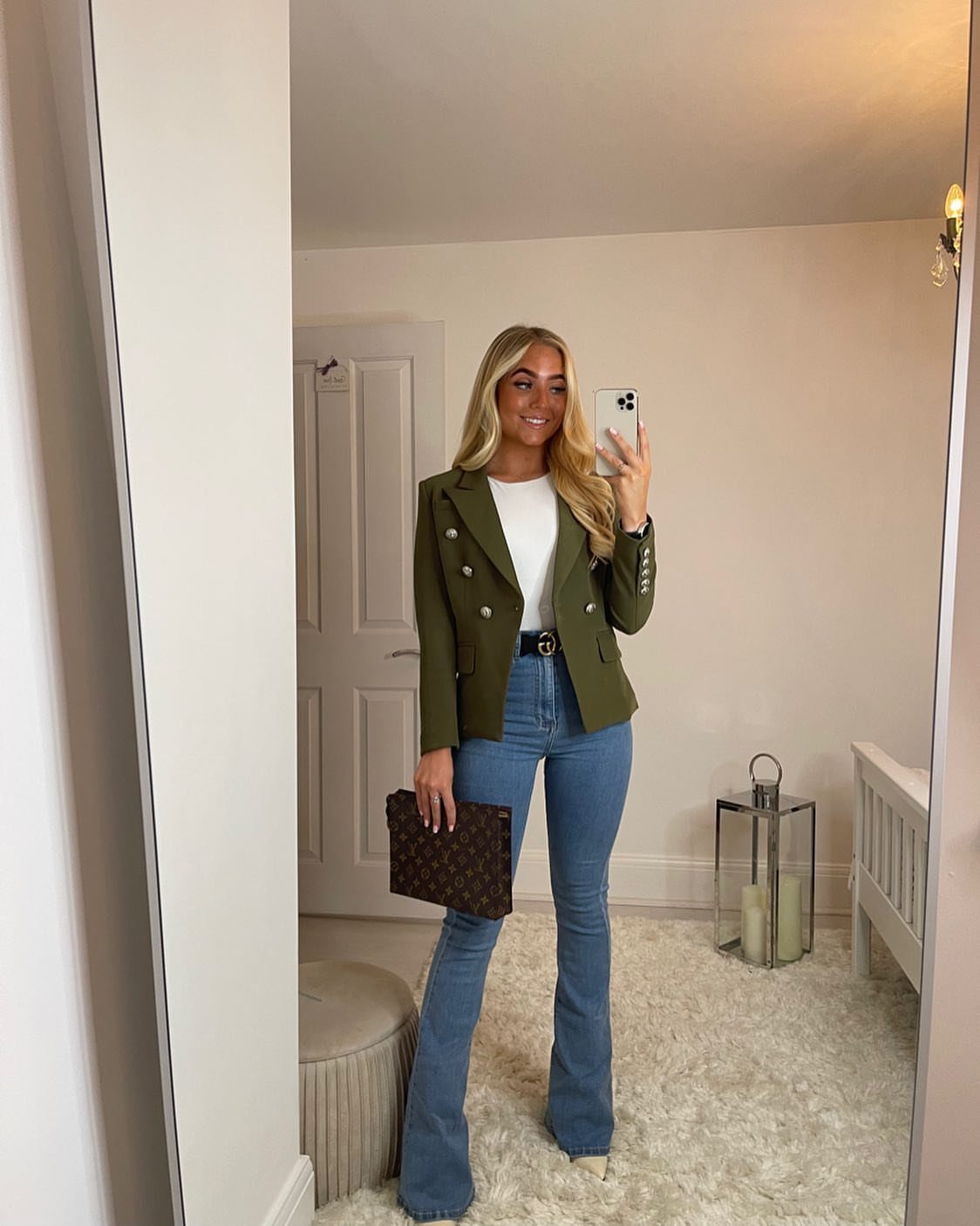Woman posing in a mirror wearing an olive blazer, white top, and blue flared jeans.