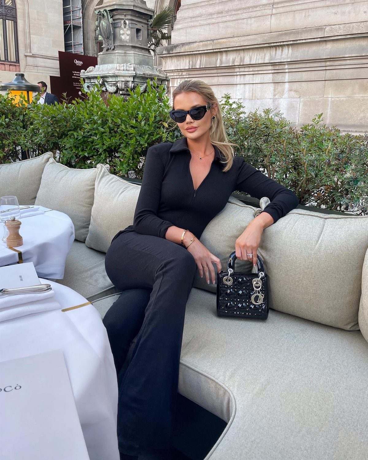 Blonde woman in a black long-sleeved jumpsuit sitting on an outdoor lounge sofa.