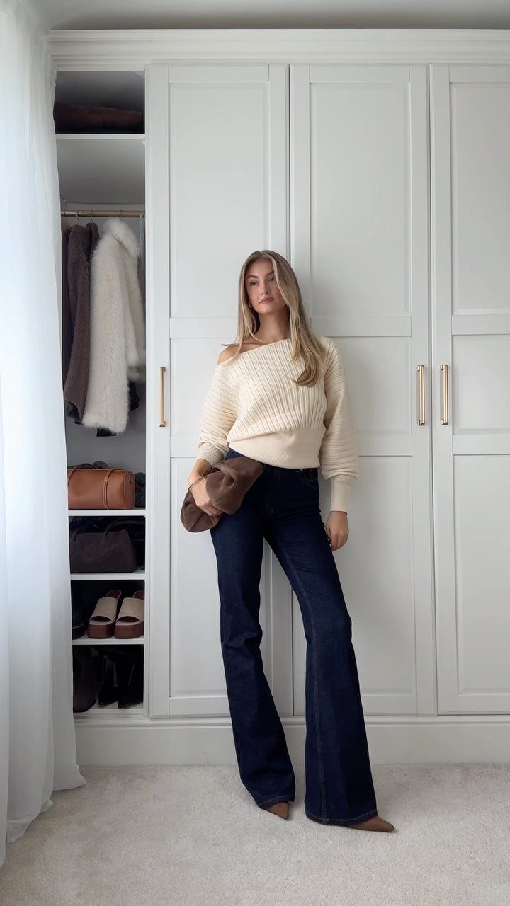 A woman posing in a cream off-the-shoulder sweater and dark blue flared jeans.