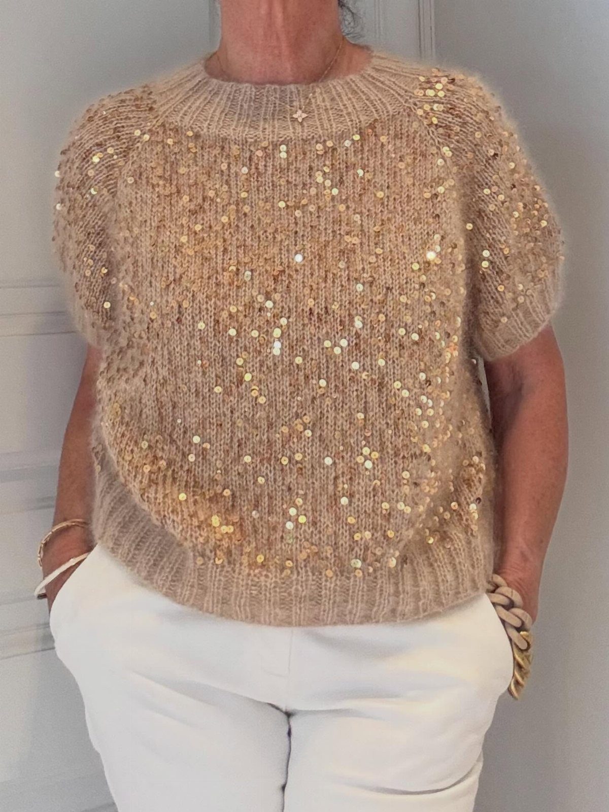 A person wearing a beige sequined knit sweater and white trousers.