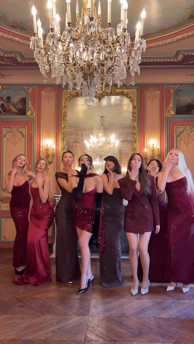 Women posing in a grand room wearing various elegant burgundy and red evening outfits.
