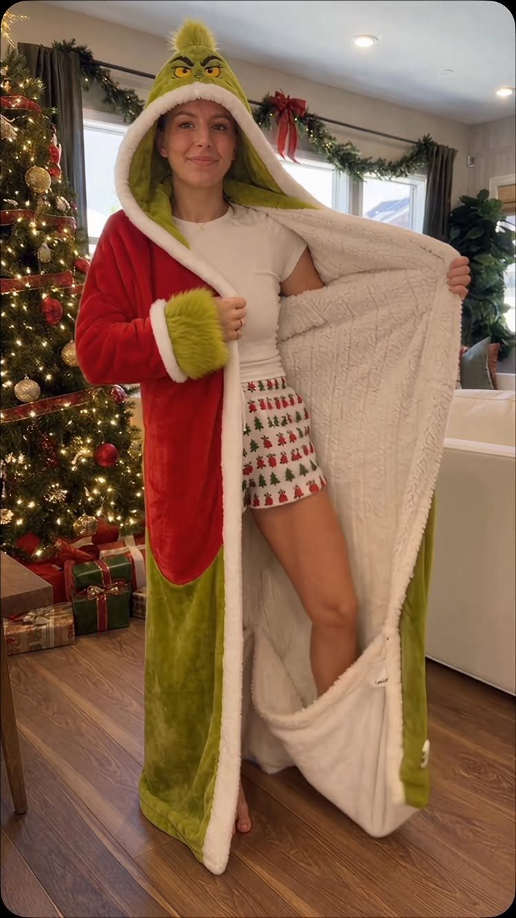 A woman wearing a red and green Grinch-themed robe and festive shorts standing near a Christmas tree.