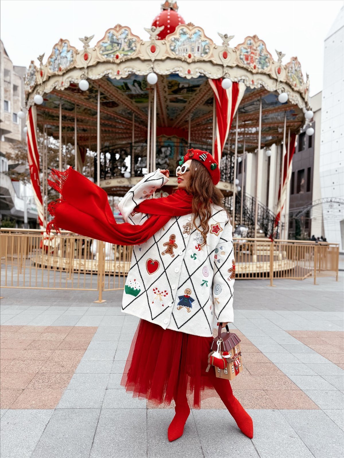 Woman in festive white patterned coat, red tulle skirt, and red scarf standing before a carousel.