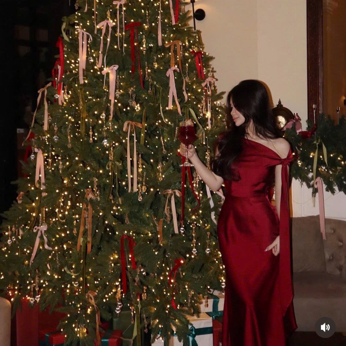 Woman in a long red silk dress standing by a lit Christmas tree holding wine.