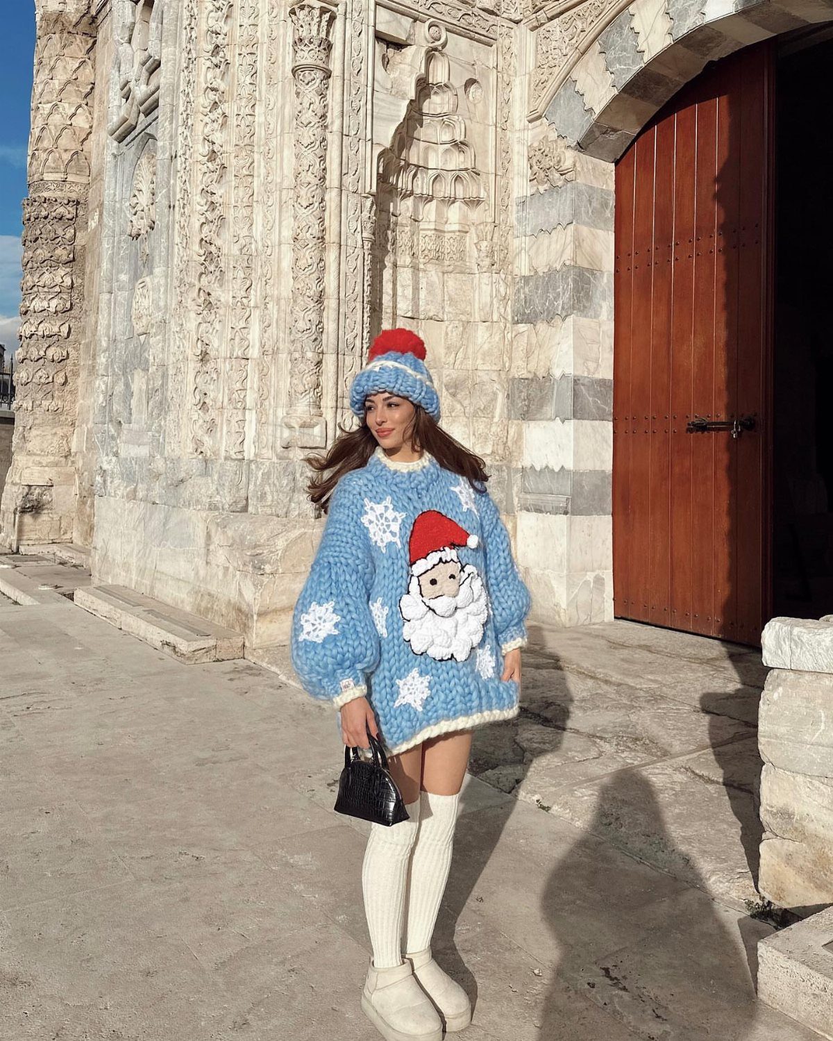 Woman wearing a blue Santa Claus sweater, matching beanie, white socks, and boots standing outdoors.